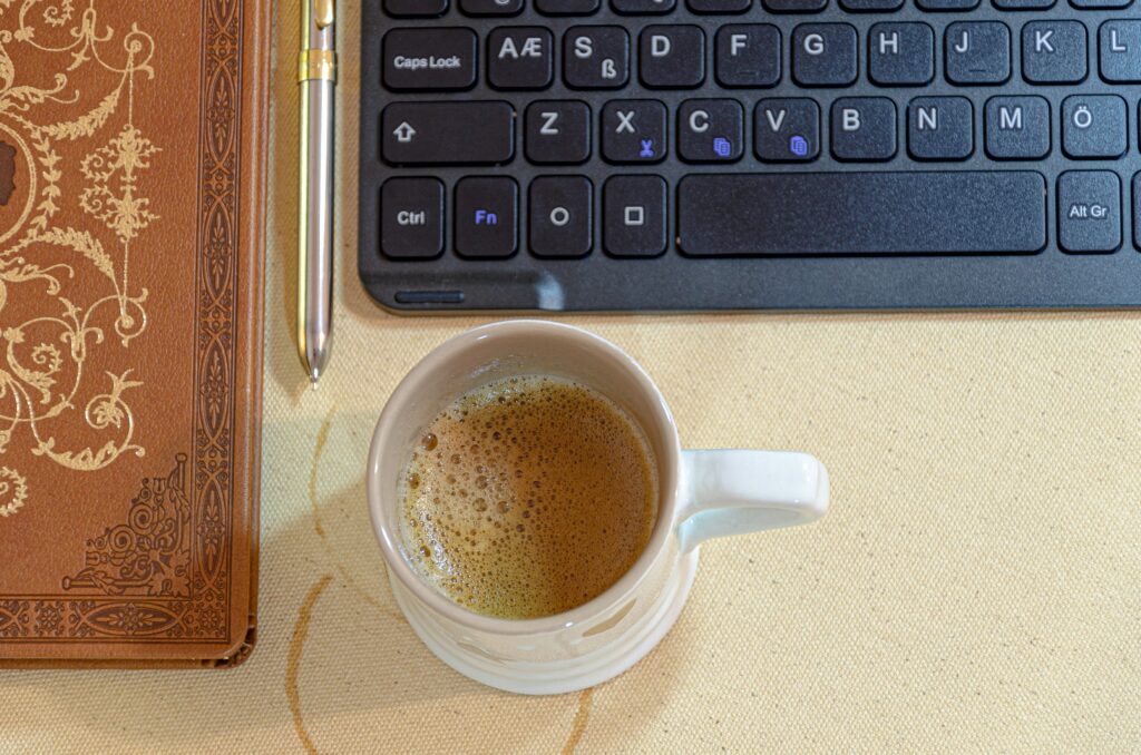 A cup of coffee beside a keyboard and a notebook