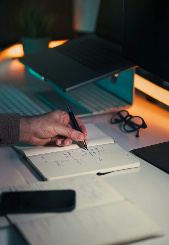 A hand writing notes in a notebook beside a pair of glasses